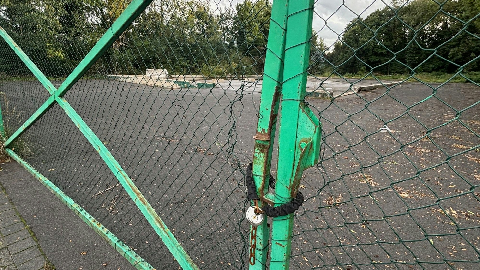 Auf dem Bild ist ein Zaun mit einem Kettenschloss vor einem brachliegenden Grundstück zu sehen.