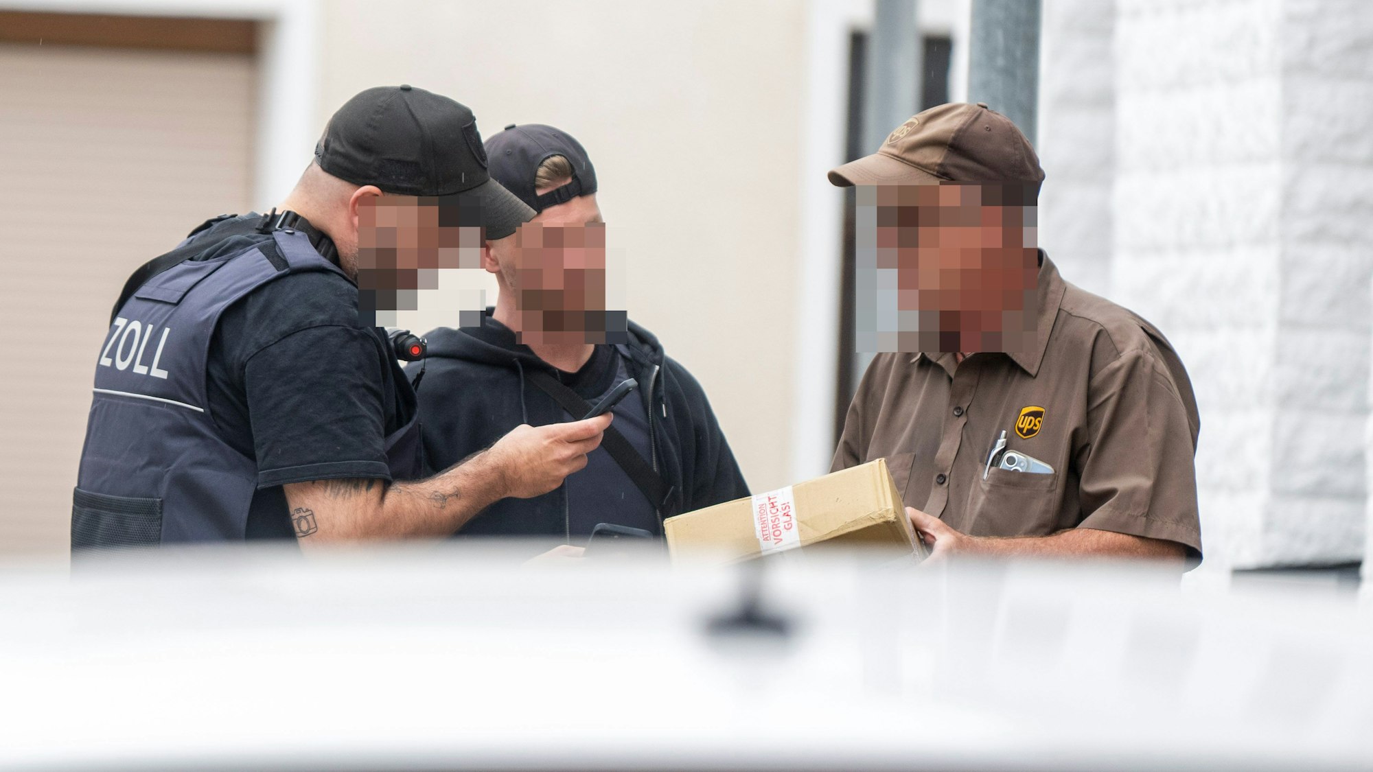 Das Bild zeigt Beamte des Zolls, die mit einem Paketzusteller sprechen und ein Foto von einem Paket machen.