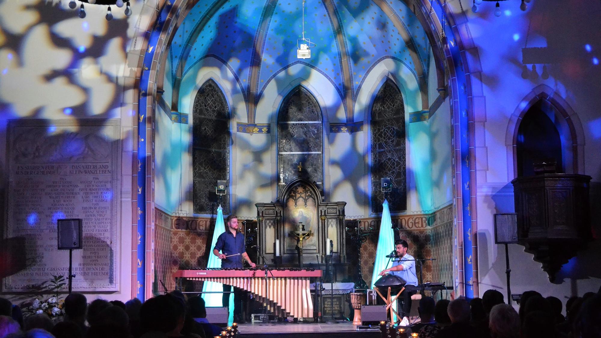 In einem in besondere Lichtprojektionen getauchtem Innenraum einer Kirche sind zwei Musiker zu sehen, einer steht am Schlagwerk, einer sitzt vor kleinen Trommeln.
