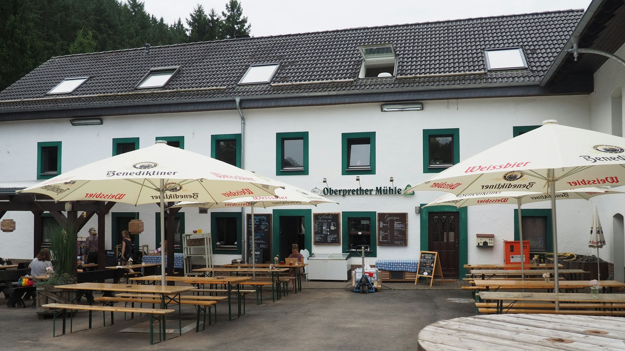 Vor dem Gebäude der Oberprether Mühle stehen Tische und Bänke unter Sonnenschirmen.