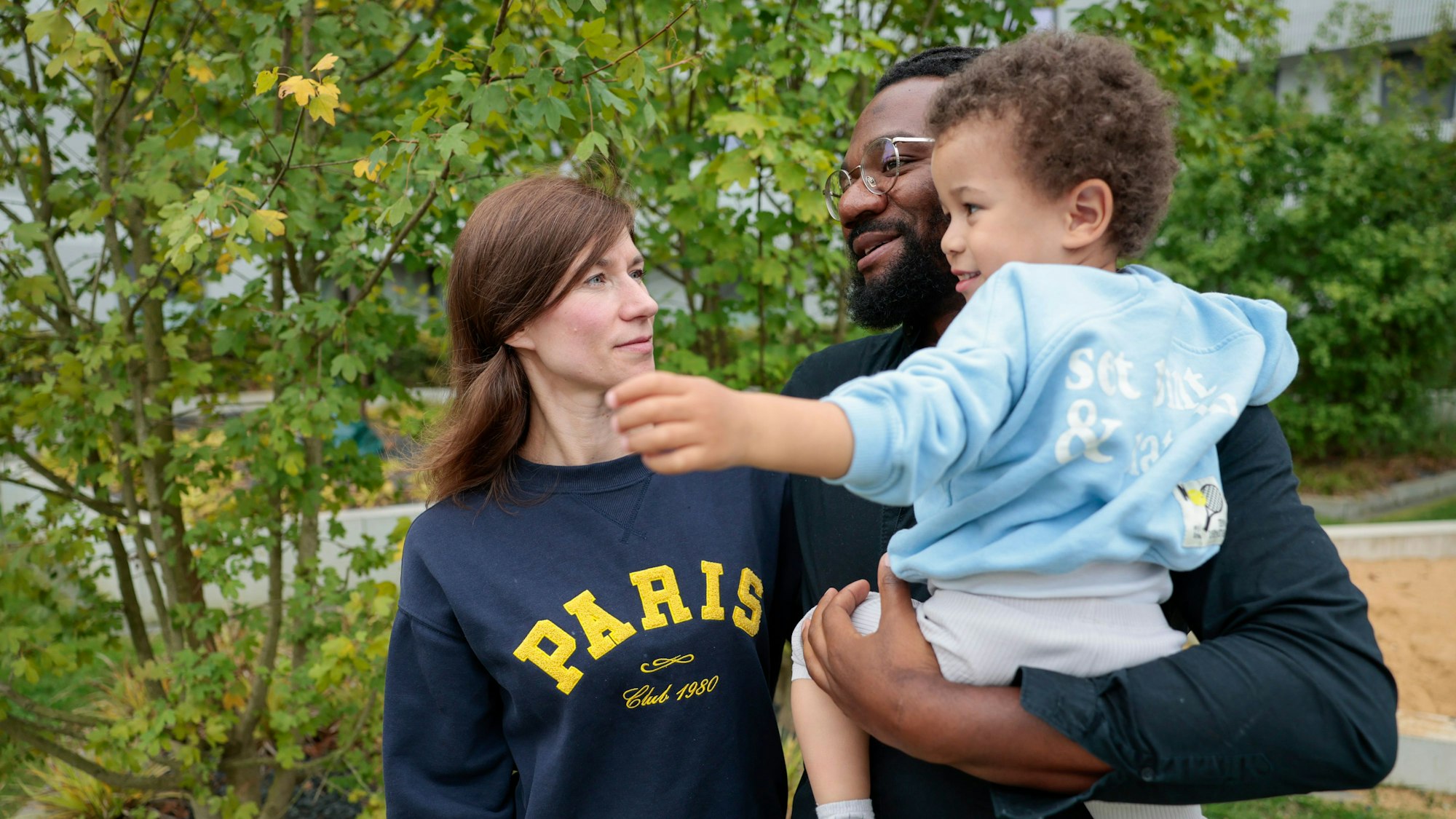 Mutter, Vater und ein fast dreijähriges Kind stehen vor einem Busch.