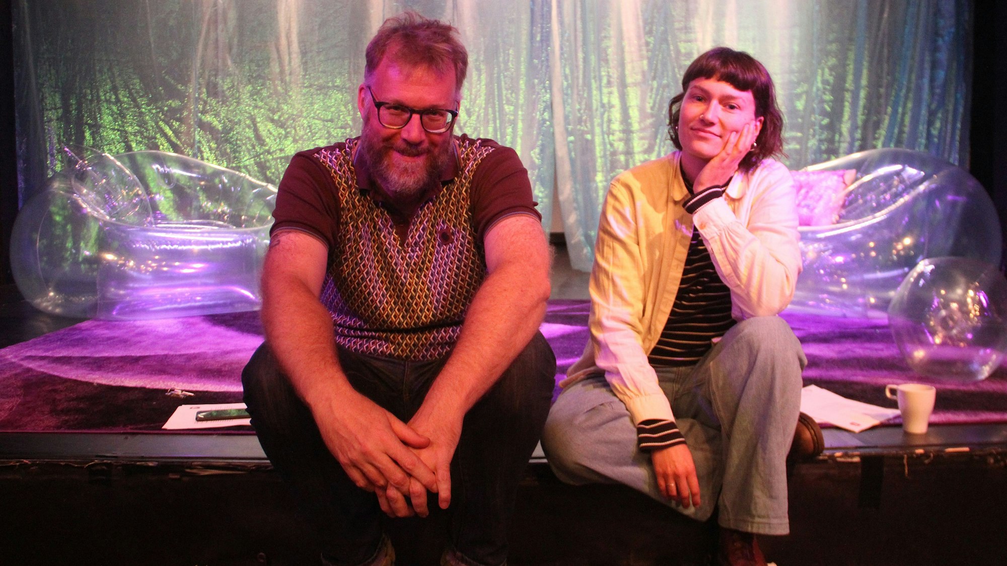 Ein Mann mit Brille und eine Frau, mit braunen Haaren, sitzen auf einem Bühnenrand.
