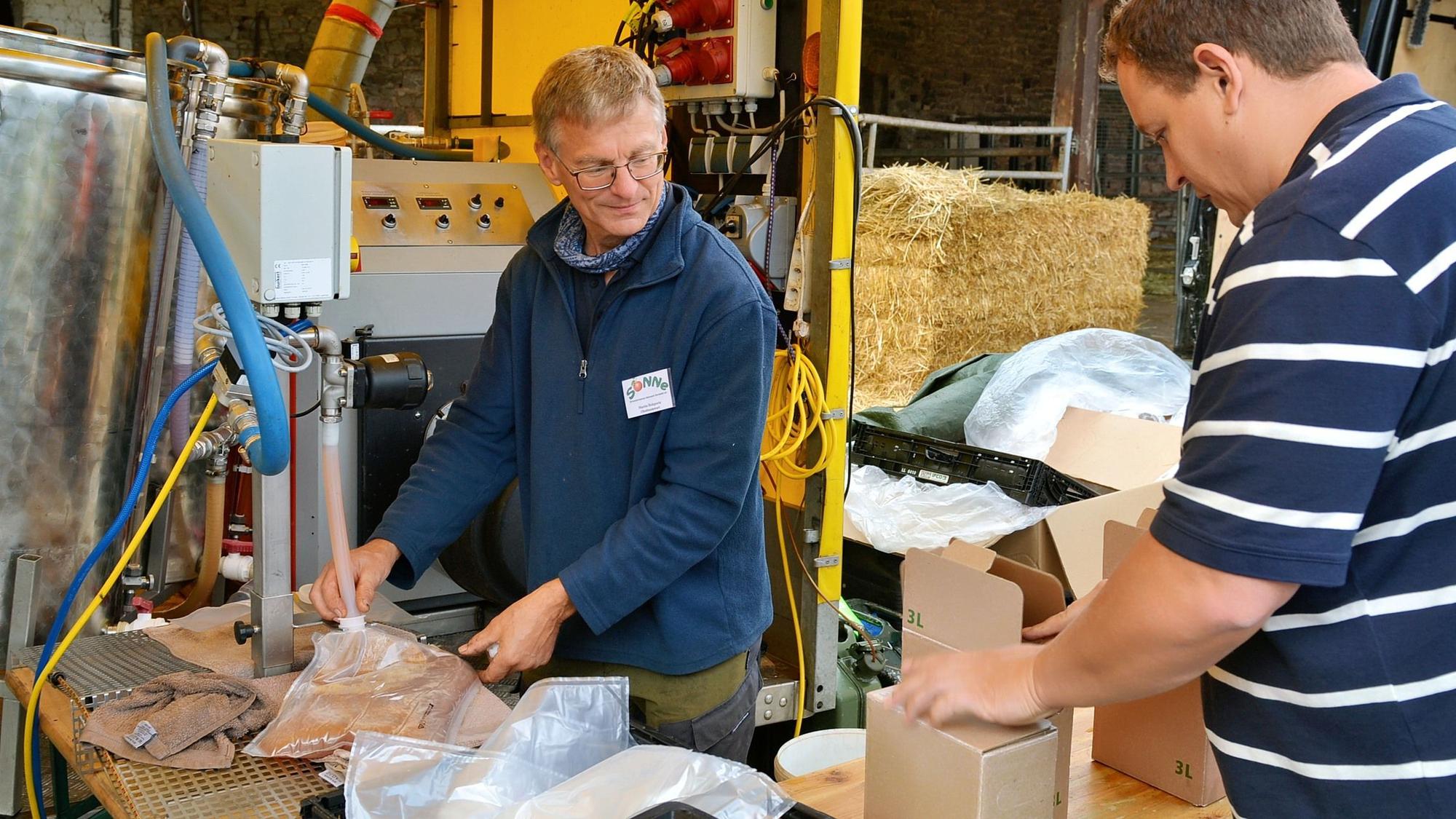 Ein Mann in einem blauen Pullover steht an einer Anlange, aus der mit einem Schlauch Apfelsaft abgefüllt wird. Ein weiterer Mann nimmt einen fertigen Karton entgegen.
