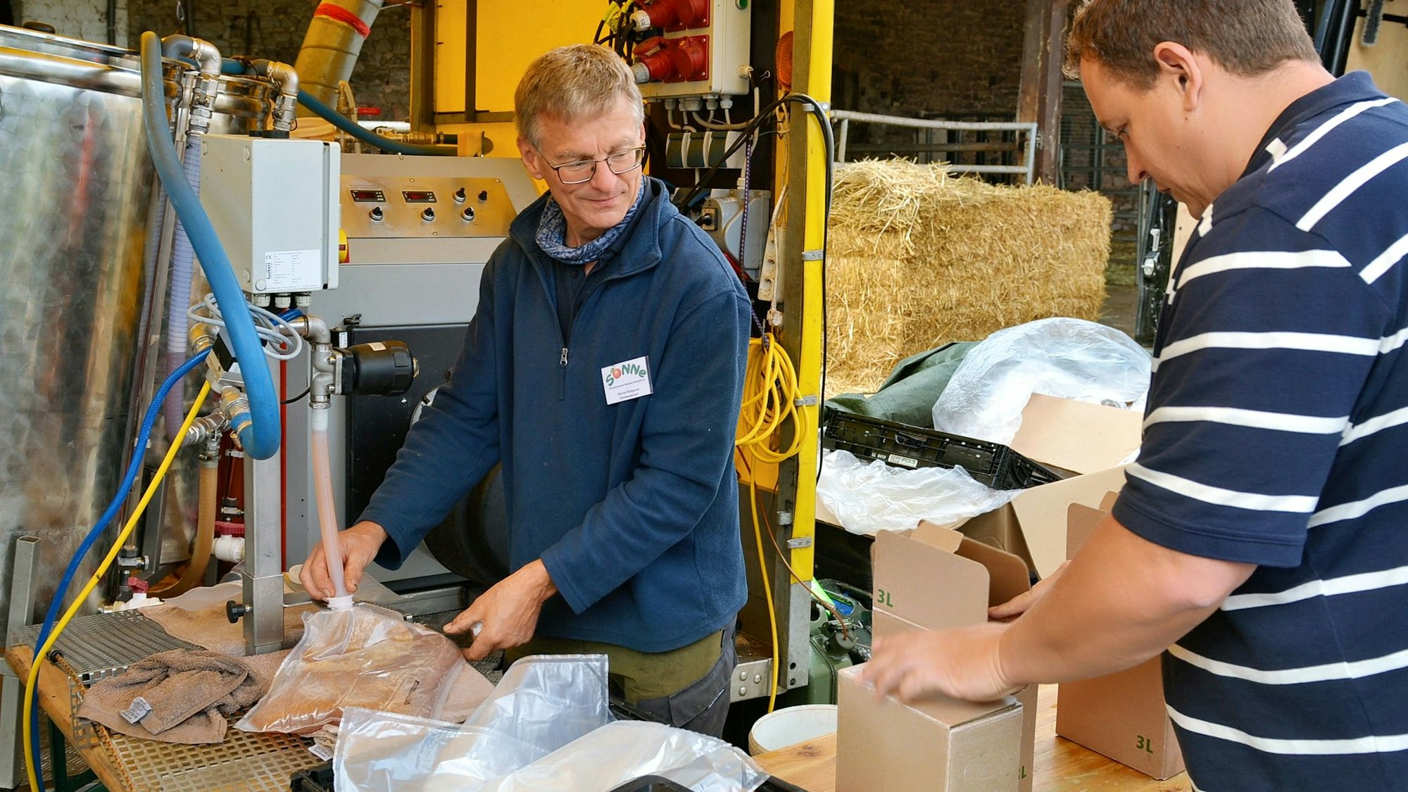 Ein Mann in einem blauen Pullover steht an einer Anlange, aus der mit einem Schlauch Apfelsaft abgefüllt wird. Ein weiterer Mann nimmt einen fertigen Karton entgegen.