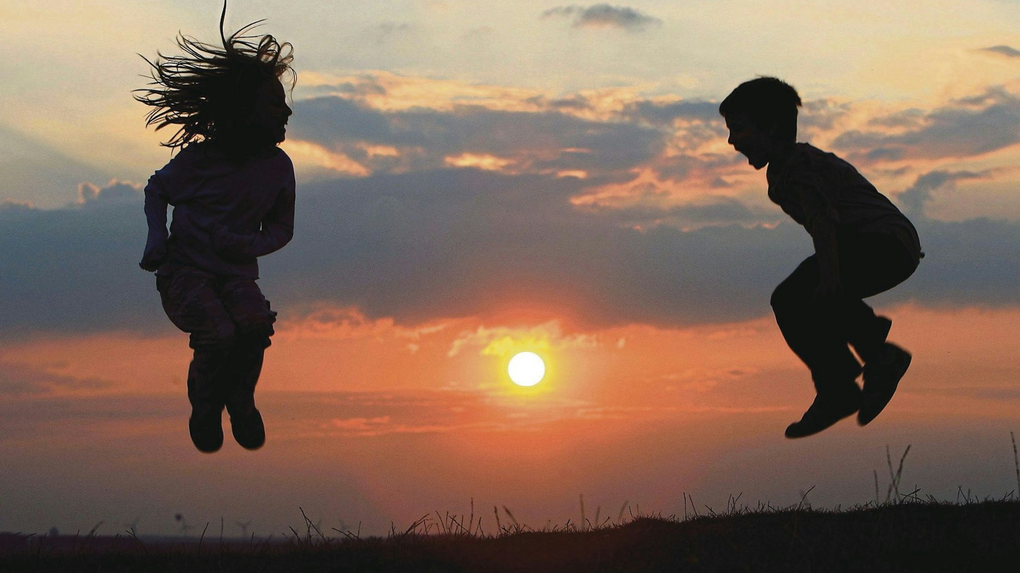 Zwei Kinder hüpfen vor einem Sonnenuntergang. Mit Entdeckungstouren durch den Landtag, Kreidebildern auf den Straßen und einem Jubiläum feiern Städte in NRW am Sonntag den Weltkindertag.