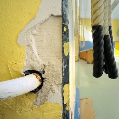 Kinder laufen in einer Turnhalle einer Schule an der Zuleitung eines Heizkörpers vorbei, in dessen Umgebung bereits der Putz abgebröckelt ist.