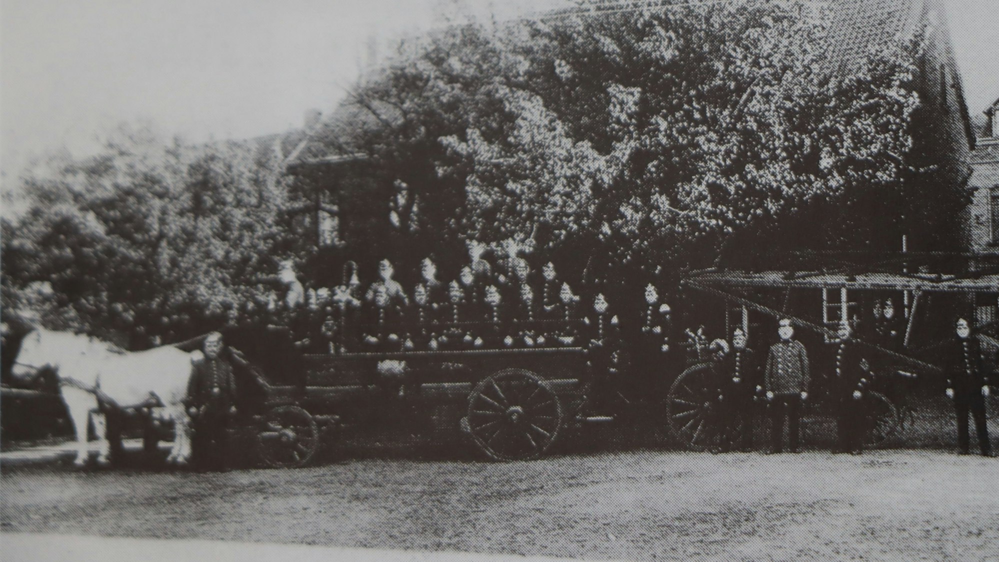 Auf dem historischen Bild sind Feuerwehrleute vor einem Wagen zu sehen, der von einem Pferd gezogen wird.