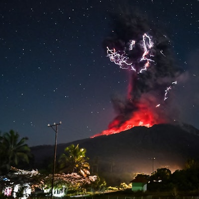 Das Bild zeigt den Kegel am Vulkan Lewotobi Laki-Lak. Bei dem Ausbruch steigt eine hohe Aschewolke in den Himmel und glühende Lava rinnt die Hänge hinunter. Durch die längere Belichtungszeit sind die Sterne am Firmament gut zu erkennen.