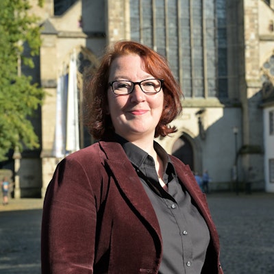 Eine Frau mit rötlichen Haaren und dunkler Brille steht vor dem Kirchenportal des Altenberger Doms.