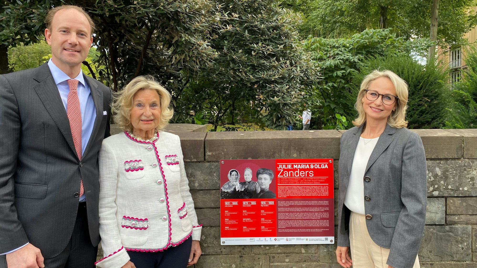 Ein Mann und zwei blonde Frauen vor einer roten Gedenktafel für drei Frauen, die in der Vergangenheit die ehemalige Papierfabrik Zanders geleitet haben.