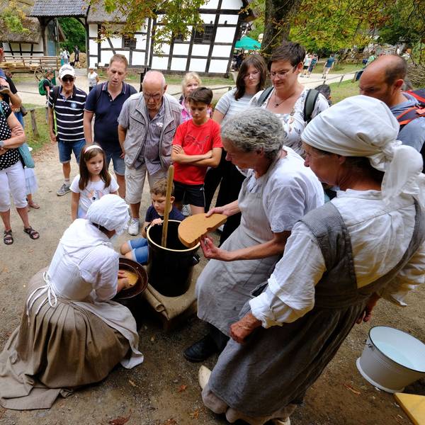 Besucher des Freilichtmuseums schauen den traditionell mit Kopftuch und Schürzen gekleideten Hauswirtschafterinnen bei ihrer Arbeit zu.