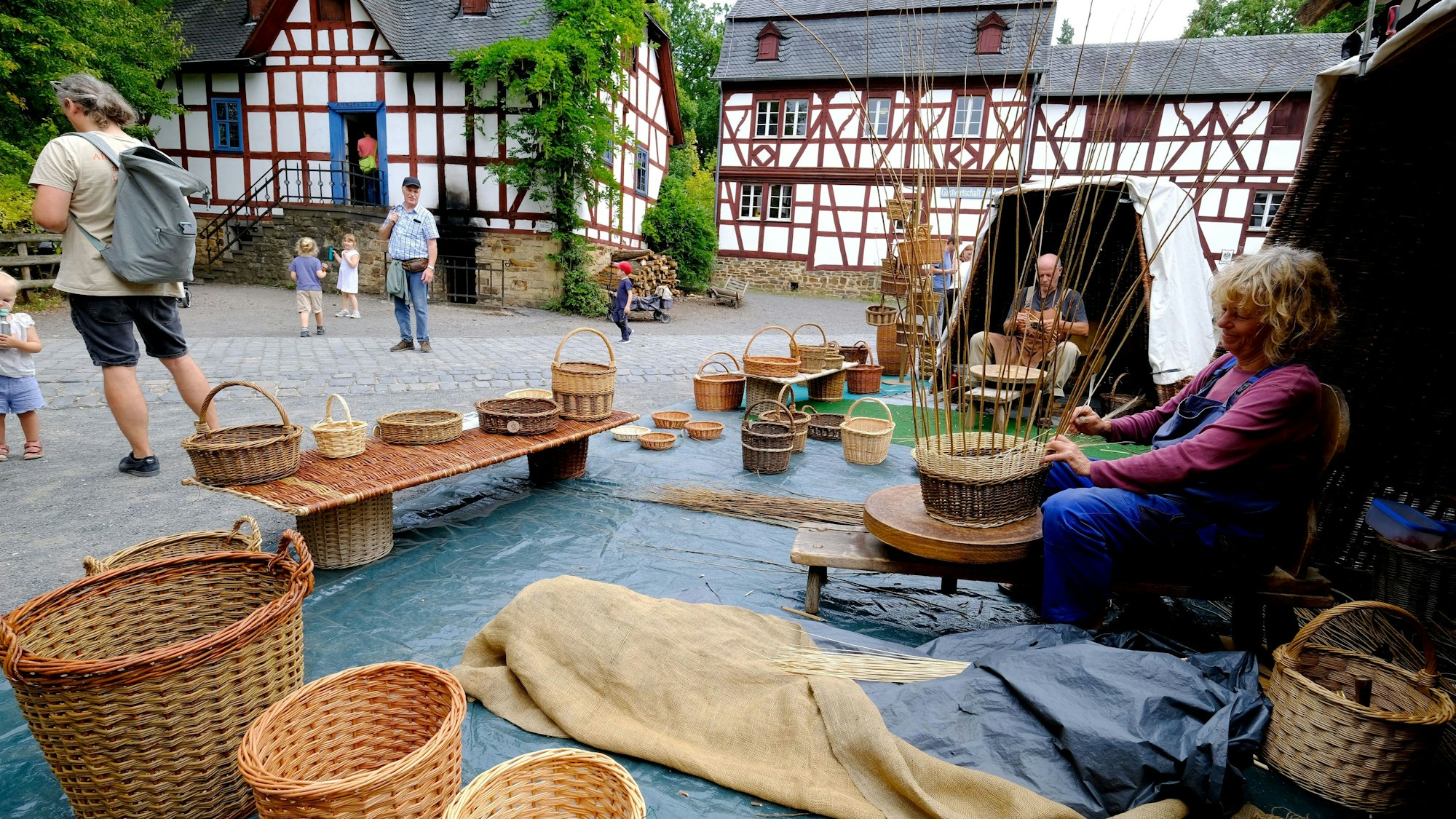 Vor den Fachwerkhäusern der Baugruppe Westerwald gehen zwei Korbmacher im Freilichtmuseum Kommern ihrem Handwerk nach.
