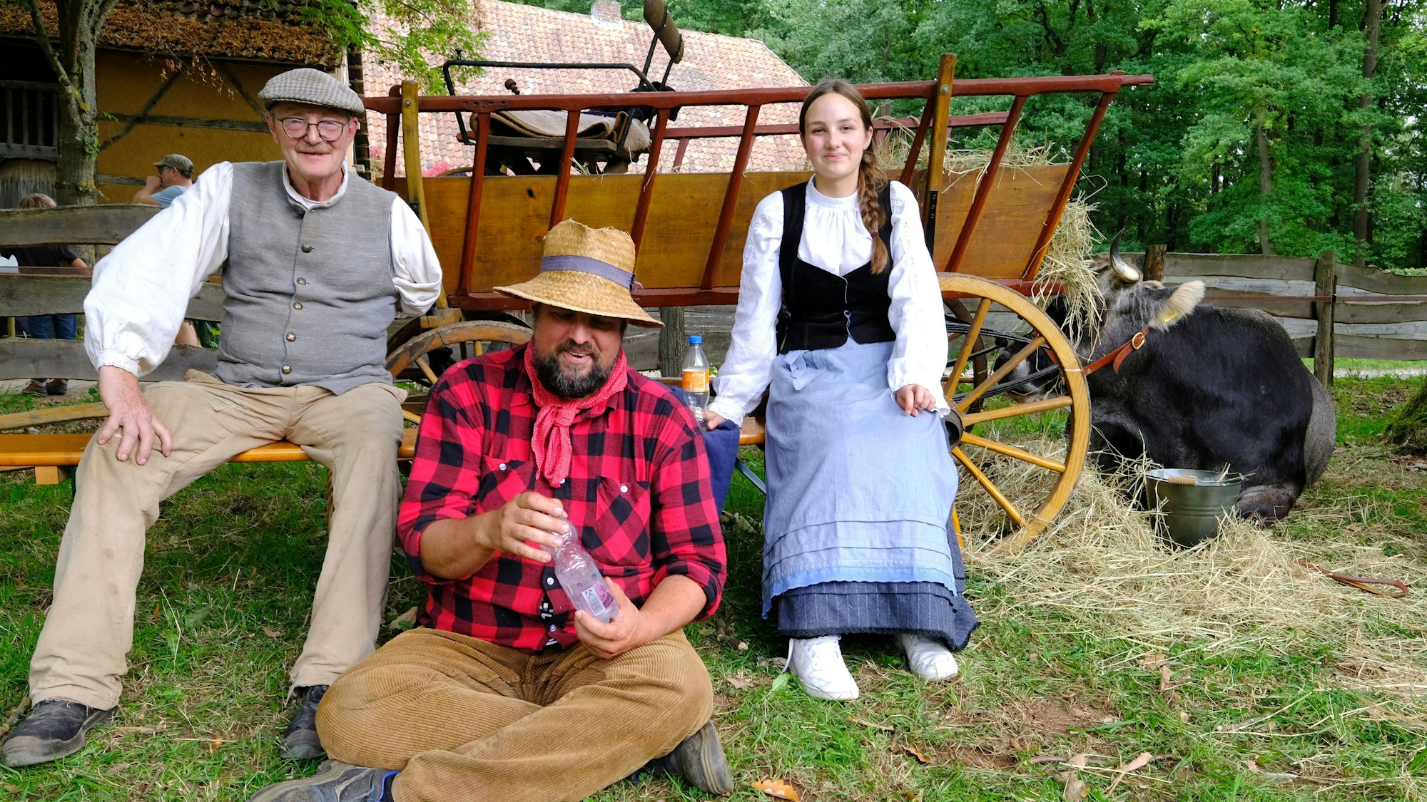 Zwei Männer und eine junge Frau sitzen vor einem Karren, neben dem eine Kuh im Gras liegt.