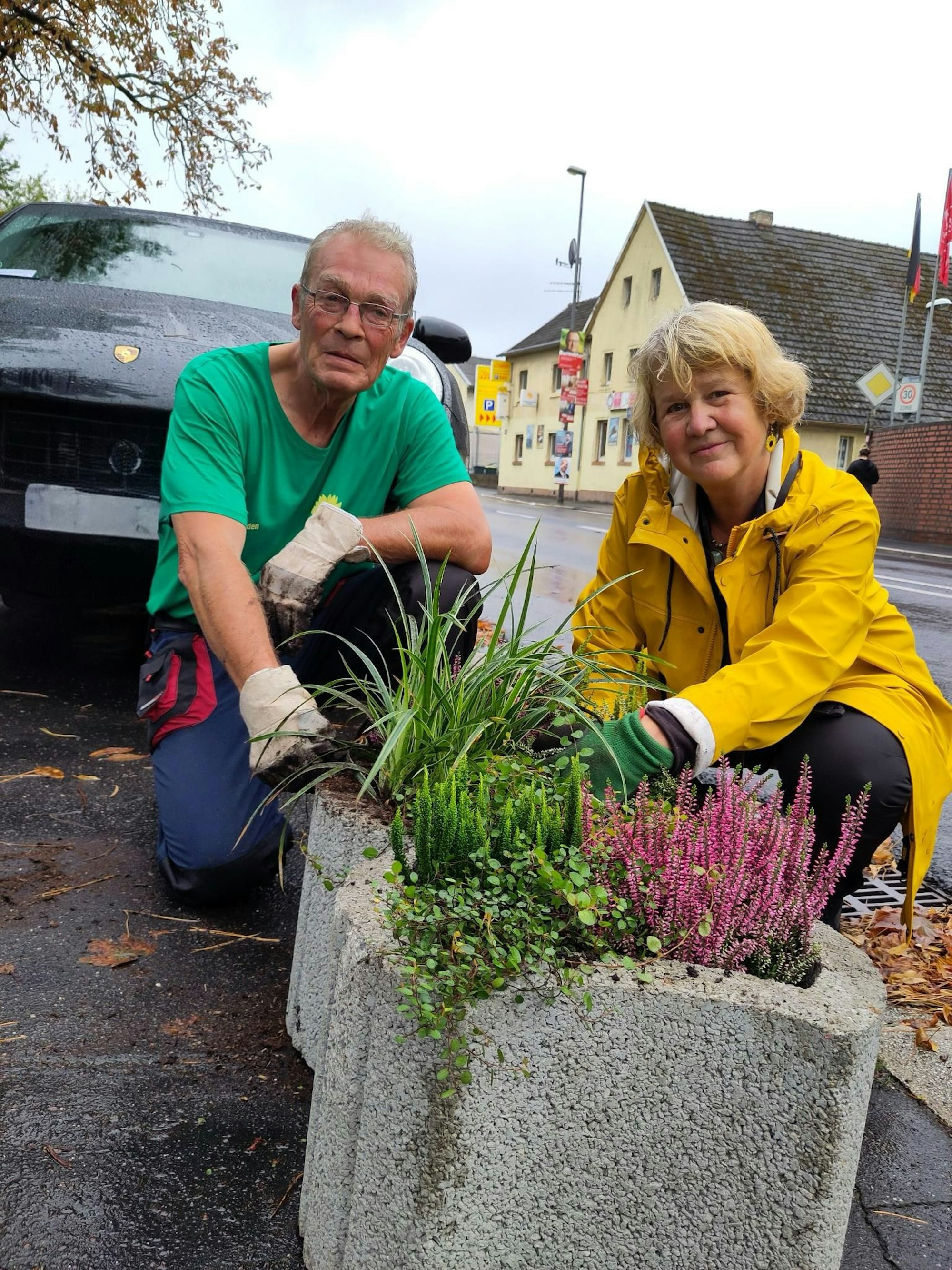 Ein Mann im grünen Shirt und eine Frau mit gelber Regenjacke hocken vor einem bepflanzten Blumenbeet an einer Straße.