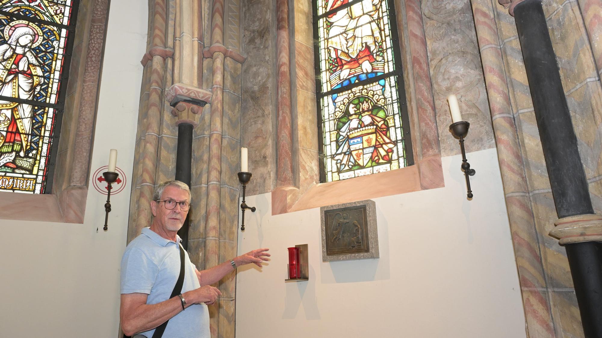 Ein grauhaariger Mann steht in einer Kapelle und zeigt auf eine Stelle der Wand.