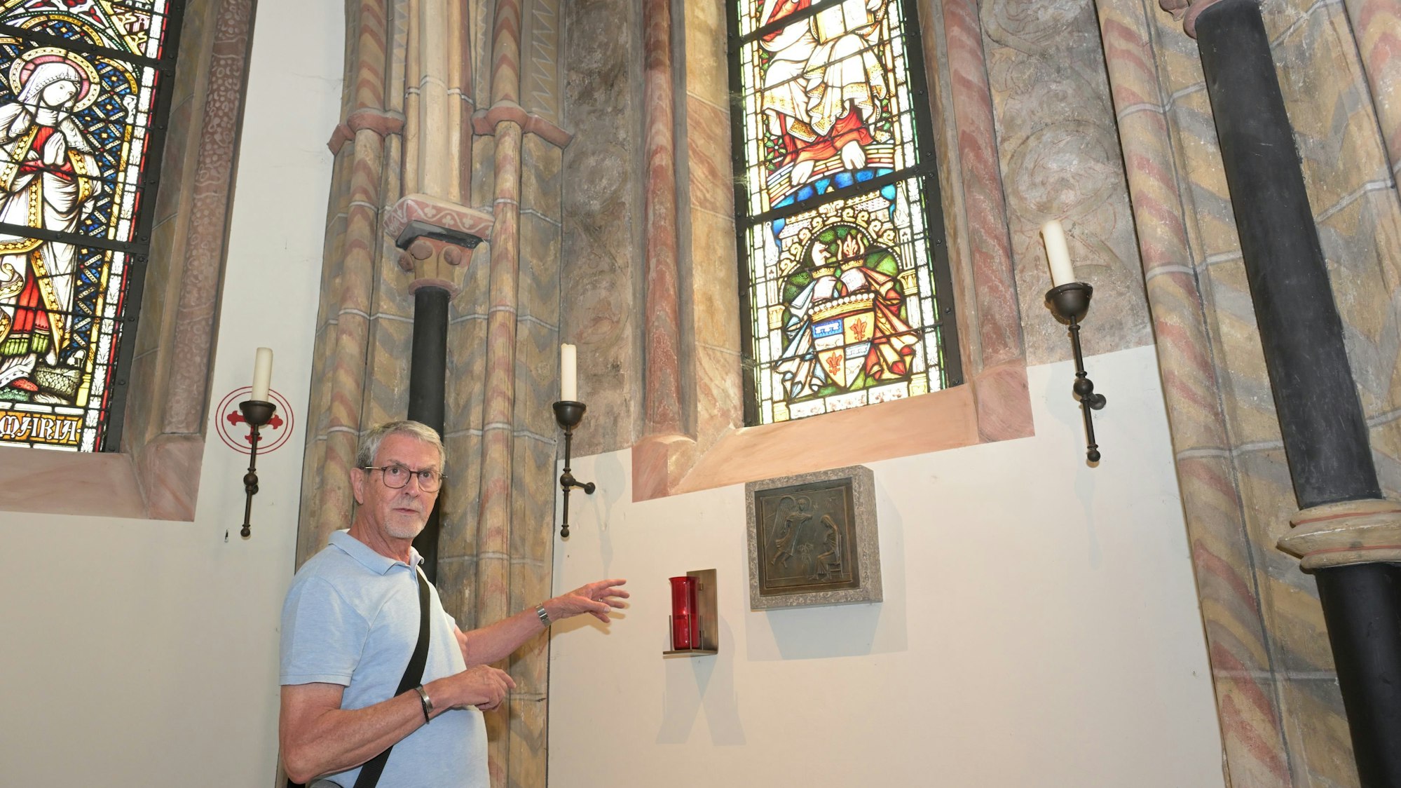 Ein grauhaariger Mann steht in einer Kapelle und zeigt auf eine Stelle der Wand.