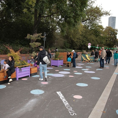 Eine Straße ist gesperrt, auf ihr stehen Sitzmöbel, sie ist mit bunten Punkten bemalt.