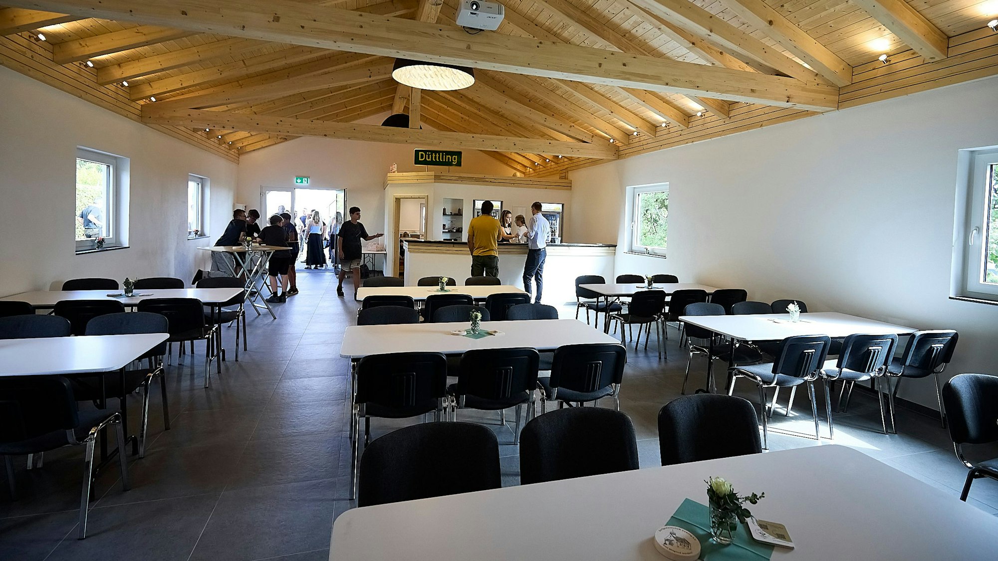 Blick in die geräumige Halle mit ihrer Holzdecke. Hinter den Tischen und Stühlen ist die Theke zu erkennen.
