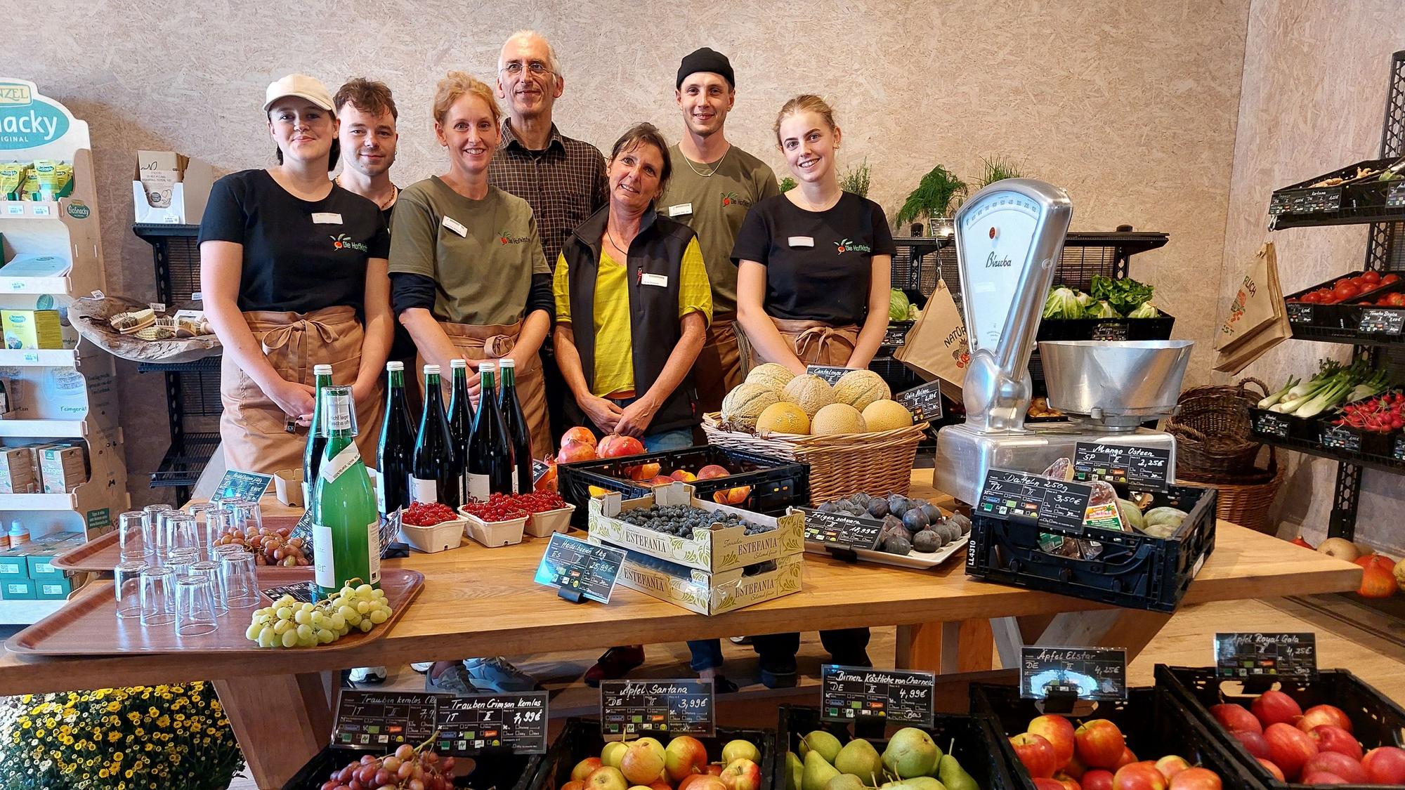 Männer und Frauen stehen hinter einem Tisch mit Obst, Gemüse und einer mechanischen Waage.