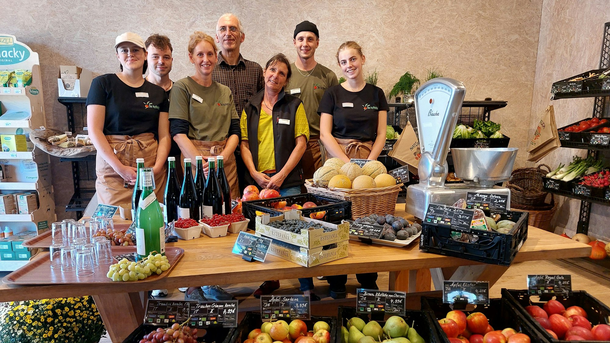 Männer und Frauen stehen hinter einem Tisch mit Obst, Gemüse und einer mechanischen Waage.