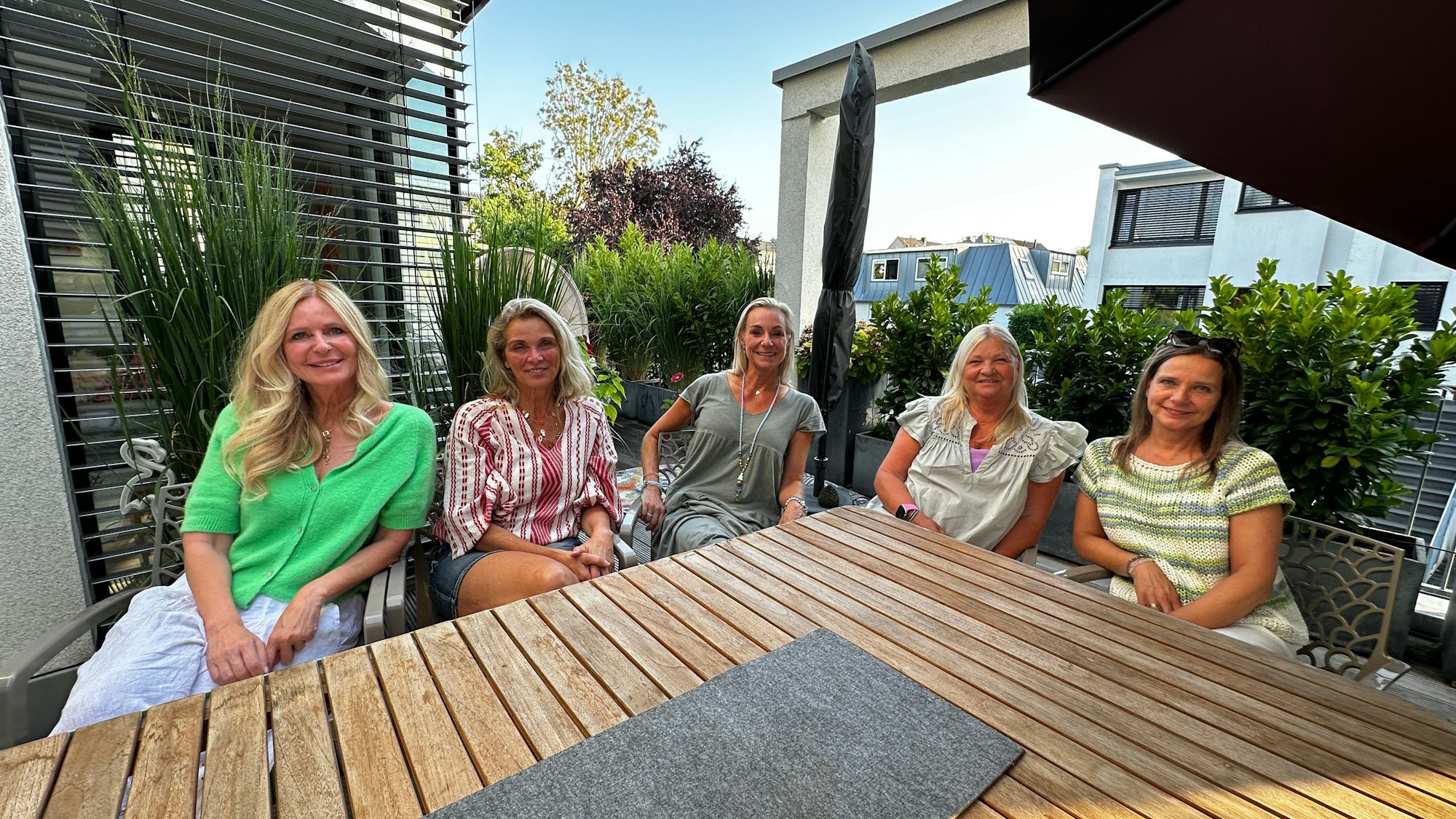 Fünf Frauen sitzen in einem Garten nebeneinander an einem Holztisch und lächeln in die Kamera.