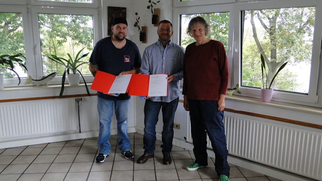 Zwei Männer und eine Frau stehen in einem Raum mit gefliesten Boden und vielen Fenstern. Die Männer halten orangefarbene Kladden mit einer Urkunde in der Hand.