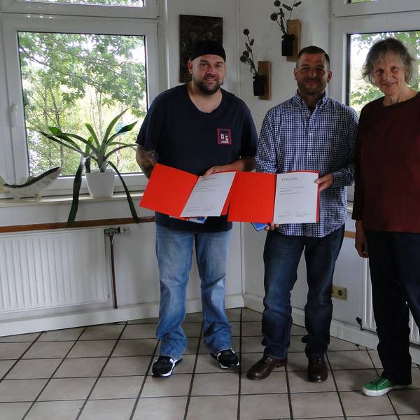 Zwei Männer und eine Frau stehen in einem Raum mit gefliesten Boden und vielen Fenstern. Die Männer halten orangefarbene Kladden mit einer Urkunde in der Hand.