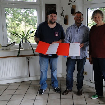 Zwei Männer und eine Frau stehen in einem Raum mit gefliesten Boden und vielen Fenstern. Die Männer halten orangefarbene Kladden mit einer Urkunde in der Hand.