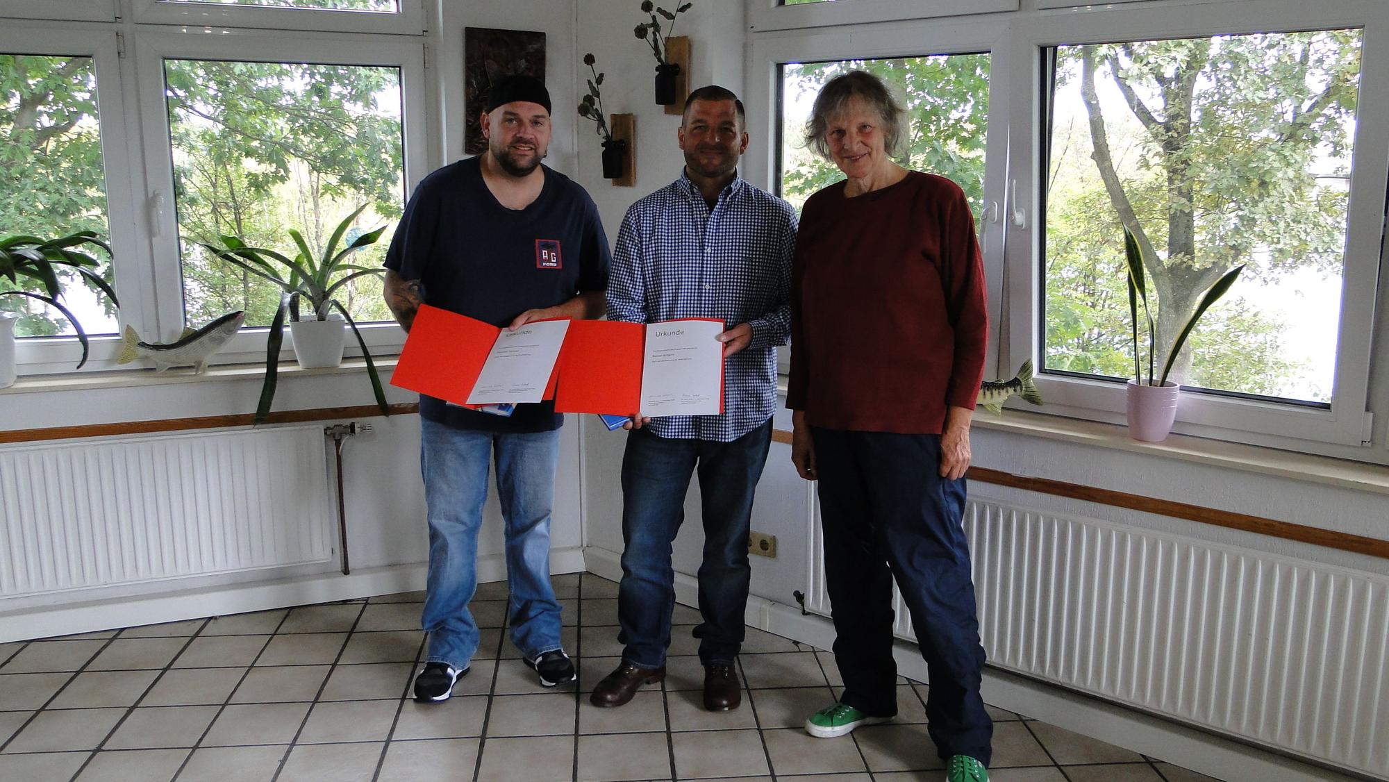 Zwei Männer und eine Frau stehen in einem Raum mit gefliesten Boden und vielen Fenstern. Die Männer halten orangefarbene Kladden mit einer Urkunde in der Hand.