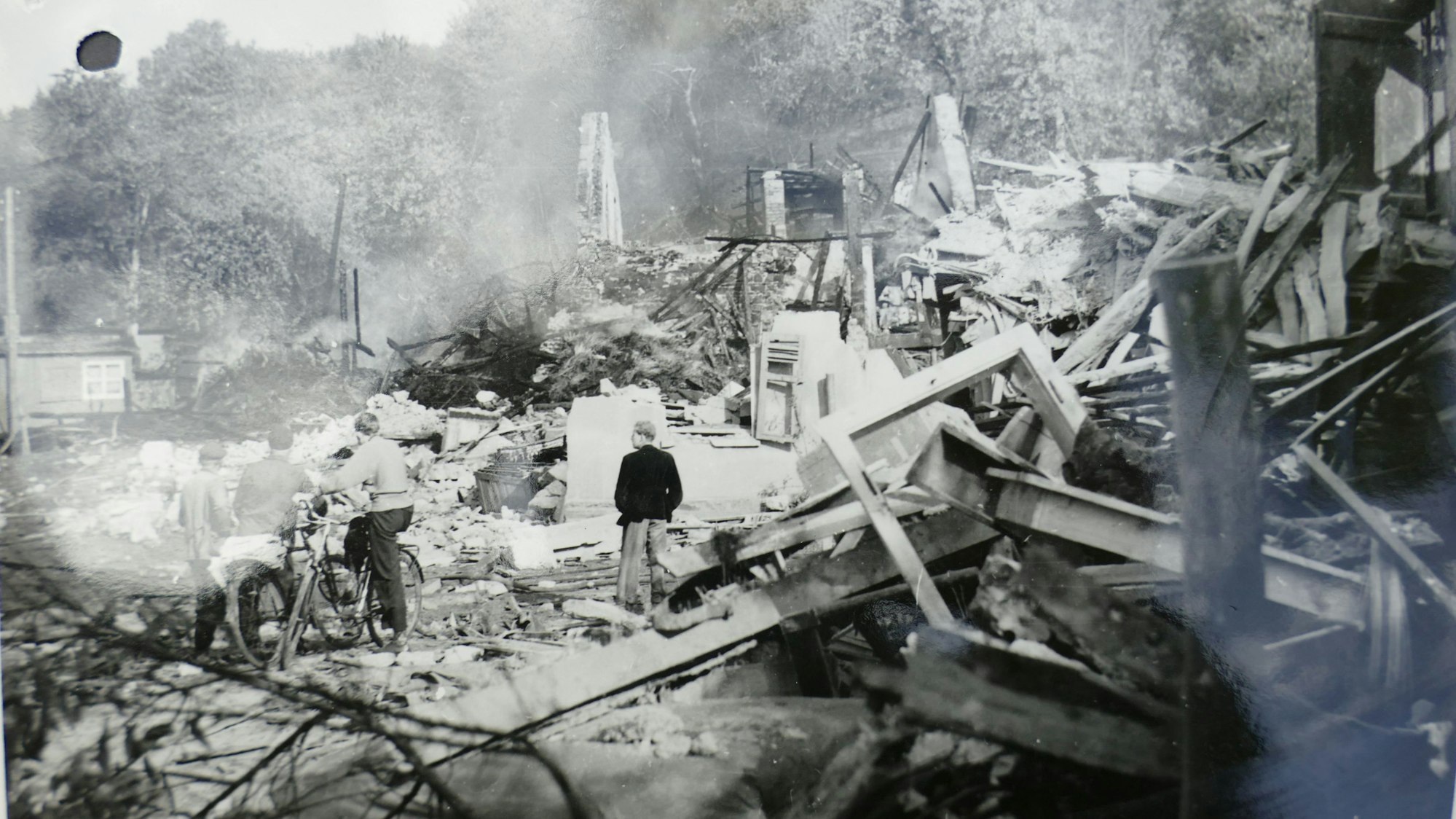 Trümmer von Häusern liegen verstreut. Mittendrin stehen Menschen, die sich das Chaos ansehen.