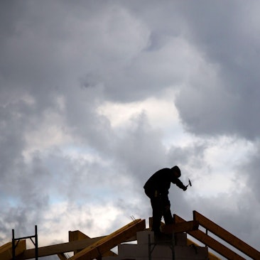 Ein Zimmermann hämmert am Dachstuhl eines im Bau befindlichen Einfamilienhauses, während im Hintergrund graue Regenwolken am Himmel zu sehen sind.