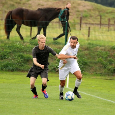 Ein ballführender Fußballer wird von einem Gegner verfolgt. Im Hintergrund führt eine Reiterin ihr Pferd.