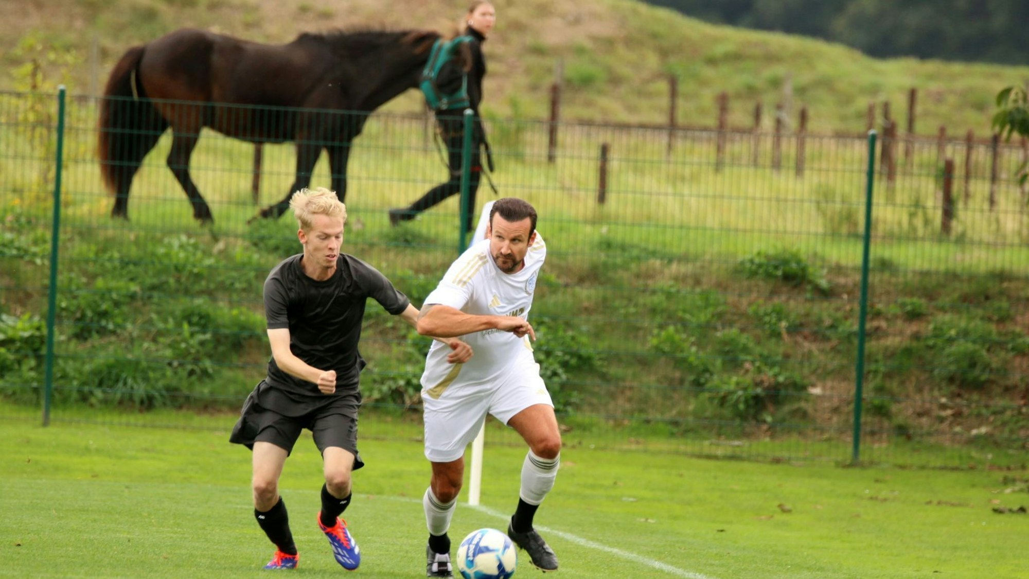 Ein ballführender Fußballer wird von einem Gegner verfolgt. Im Hintergrund führt eine Reiterin ihr Pferd.