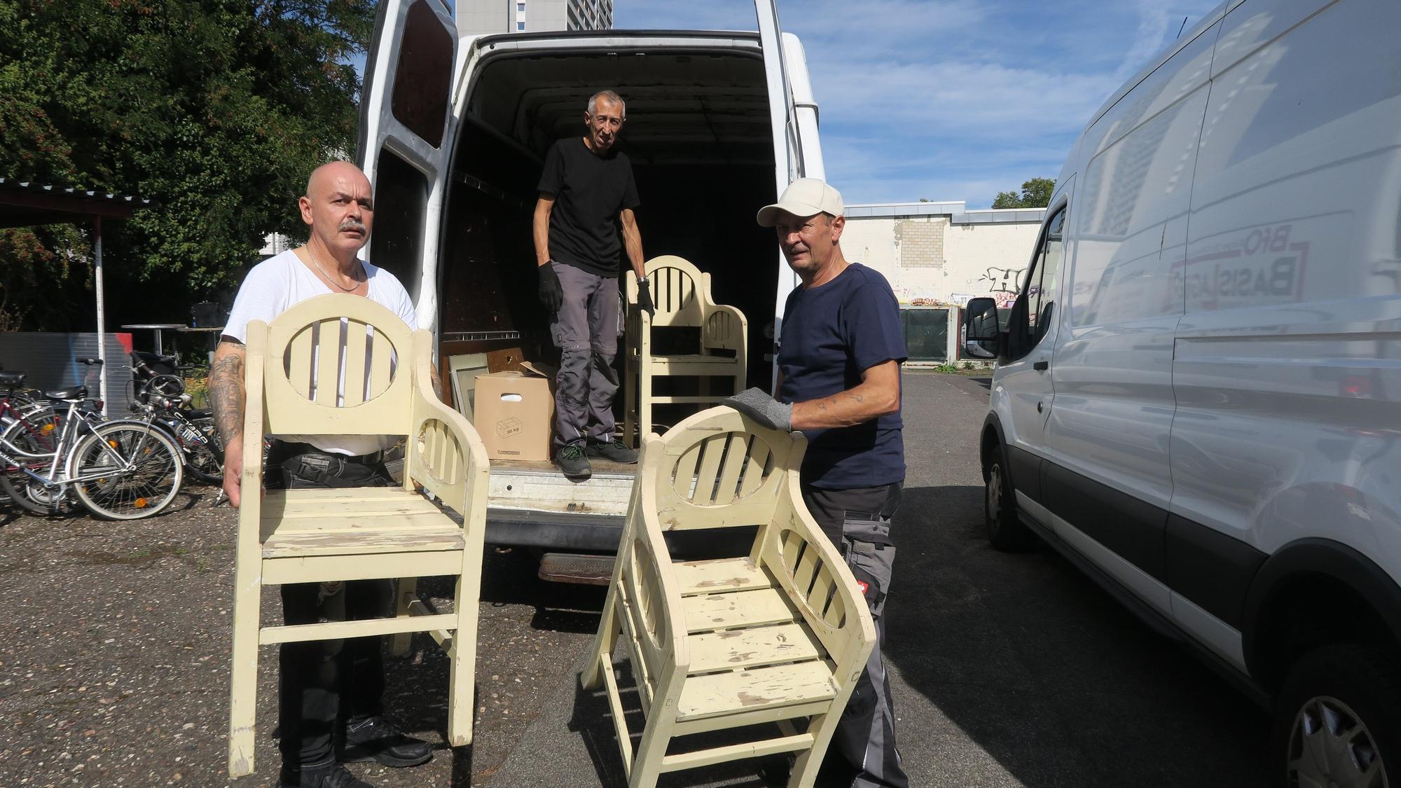 Drei Männer haben Stühle in der Hand, sie stehen neben einem Transporter.