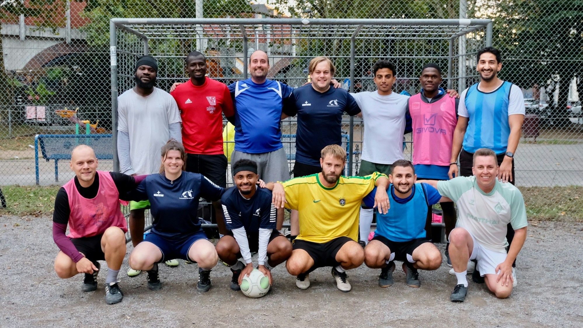 13 Personen unterschiedlicher kultureller Herkunft tragen bunte Sport-Kleidung und stehen in Form eines Mannschaftsfotos vor einem Fußballtor.