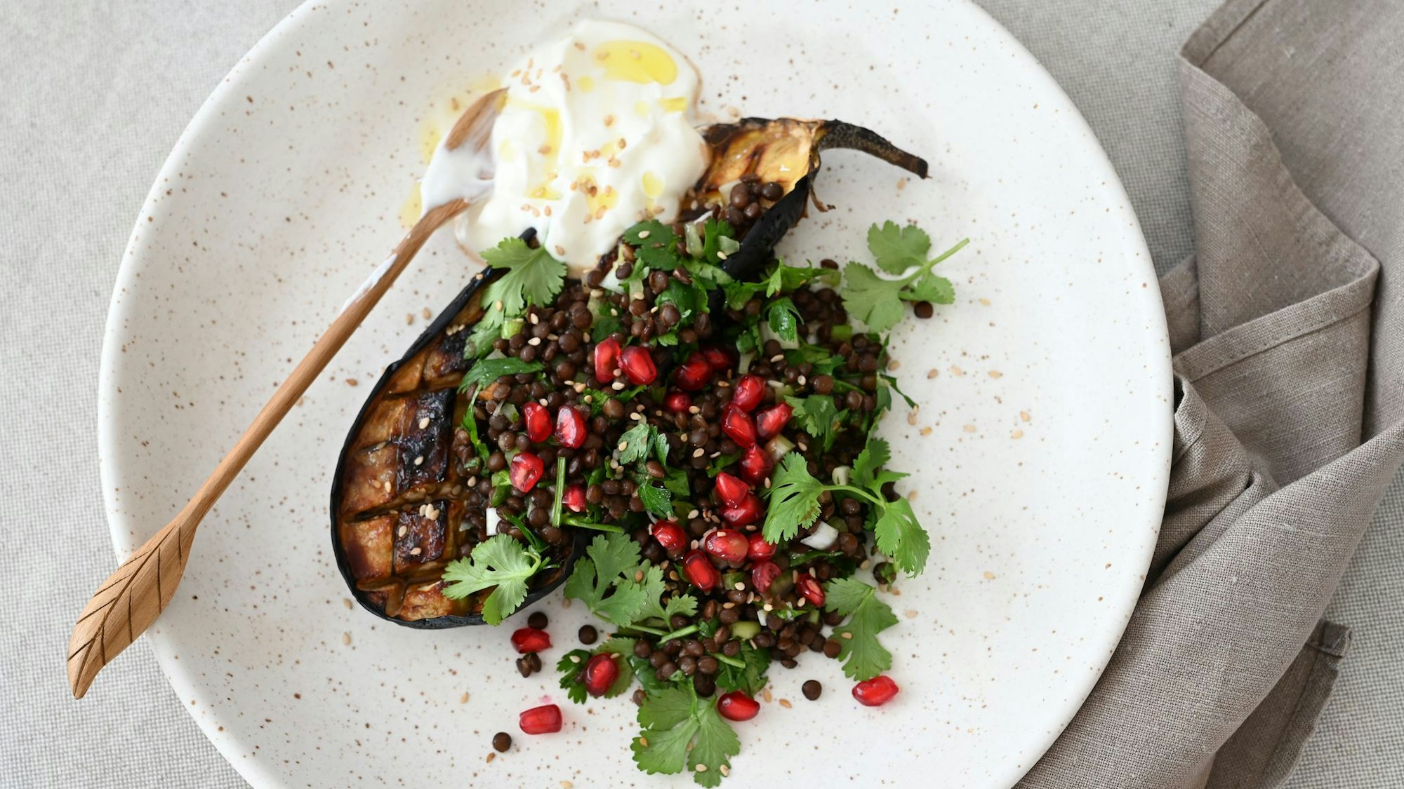Ein Teller voller Kontraste und orientalischem Twist: Geschmorte Auberginen, getoppt mit Linsen-Kräuter-Salat, Granatapfelkernen und einem Klecks cremigem Joghurt.