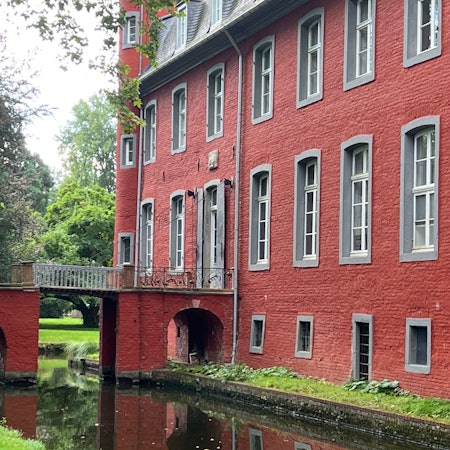 Zu sehen ist ein rotes Schloss mit einer Brücke über einen Schlossgraben.