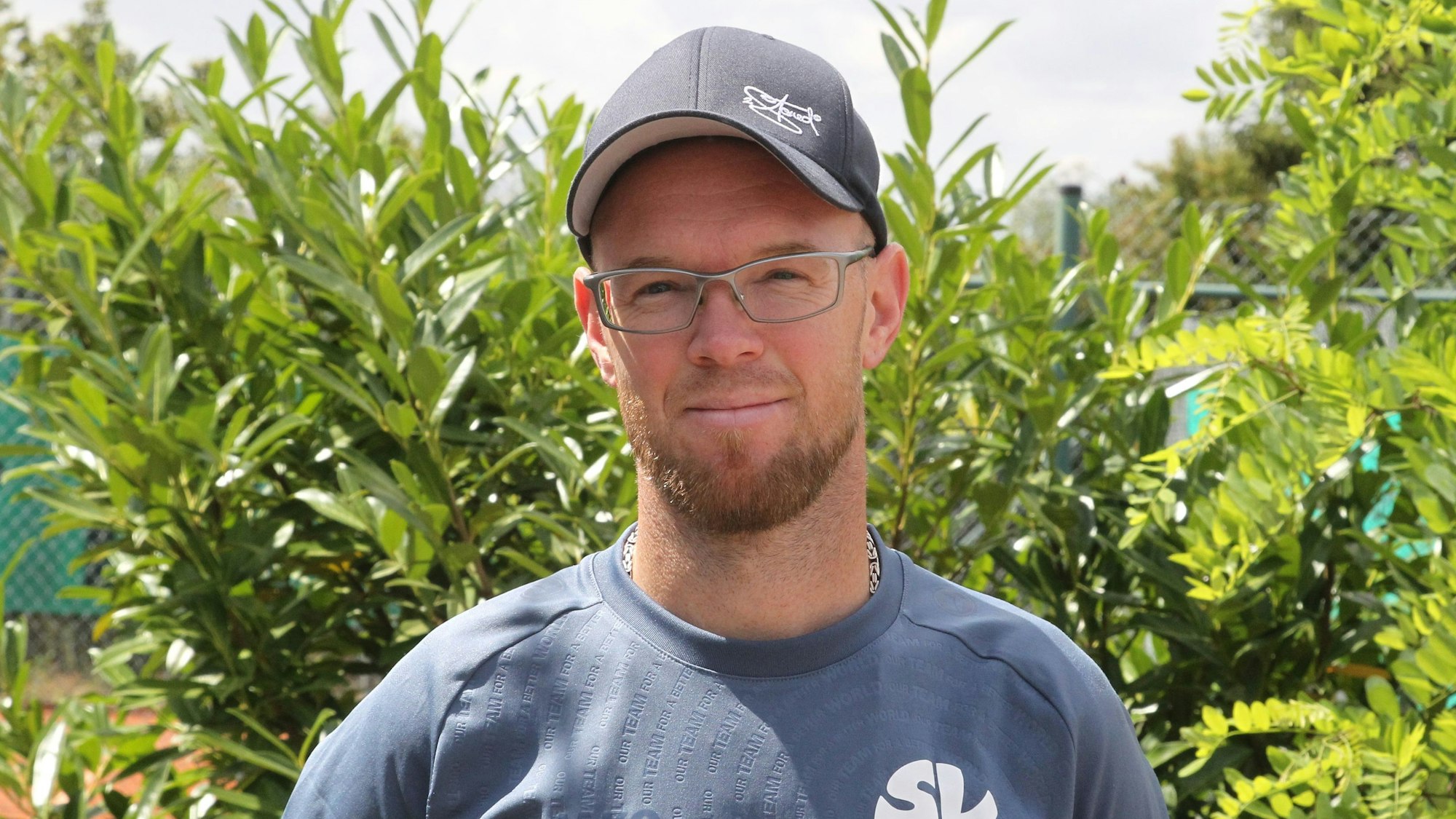 Ein Mann mit Kappe steht vor einem begrünten Hintergrund.