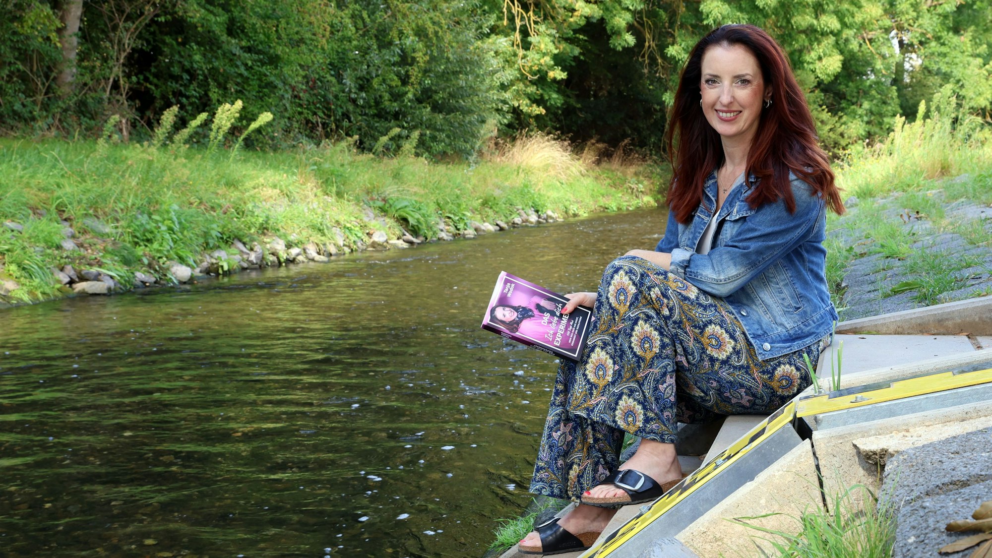 Tanja Wodok sitzt am Erftpegel bei Weilerswist und hält ihr Buch in der Hand.