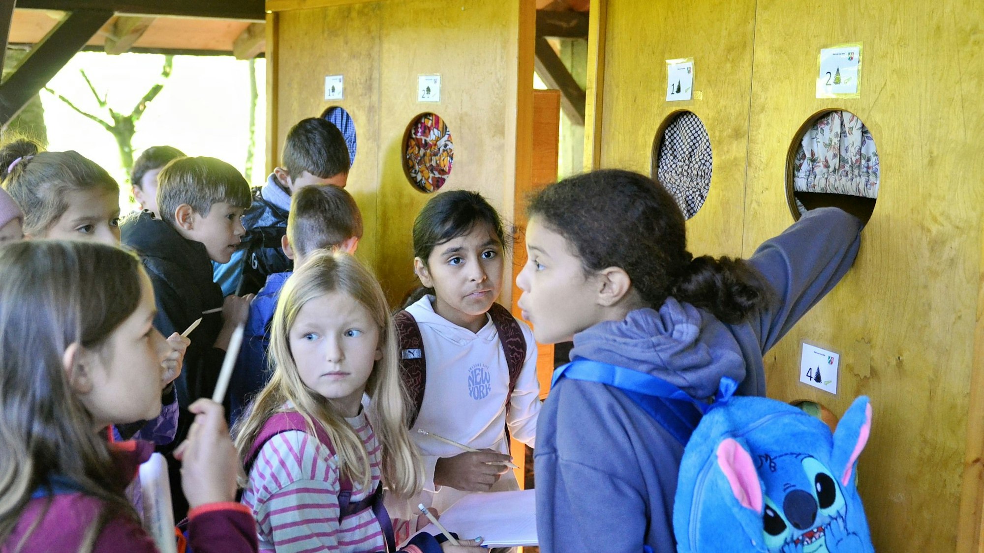 Schülerinnen und Schüler stehen vor einer Holzwand. Sie greifen in kreisrunde Öffnungen, um zu ertasten, was sich dahinter verbirgt.