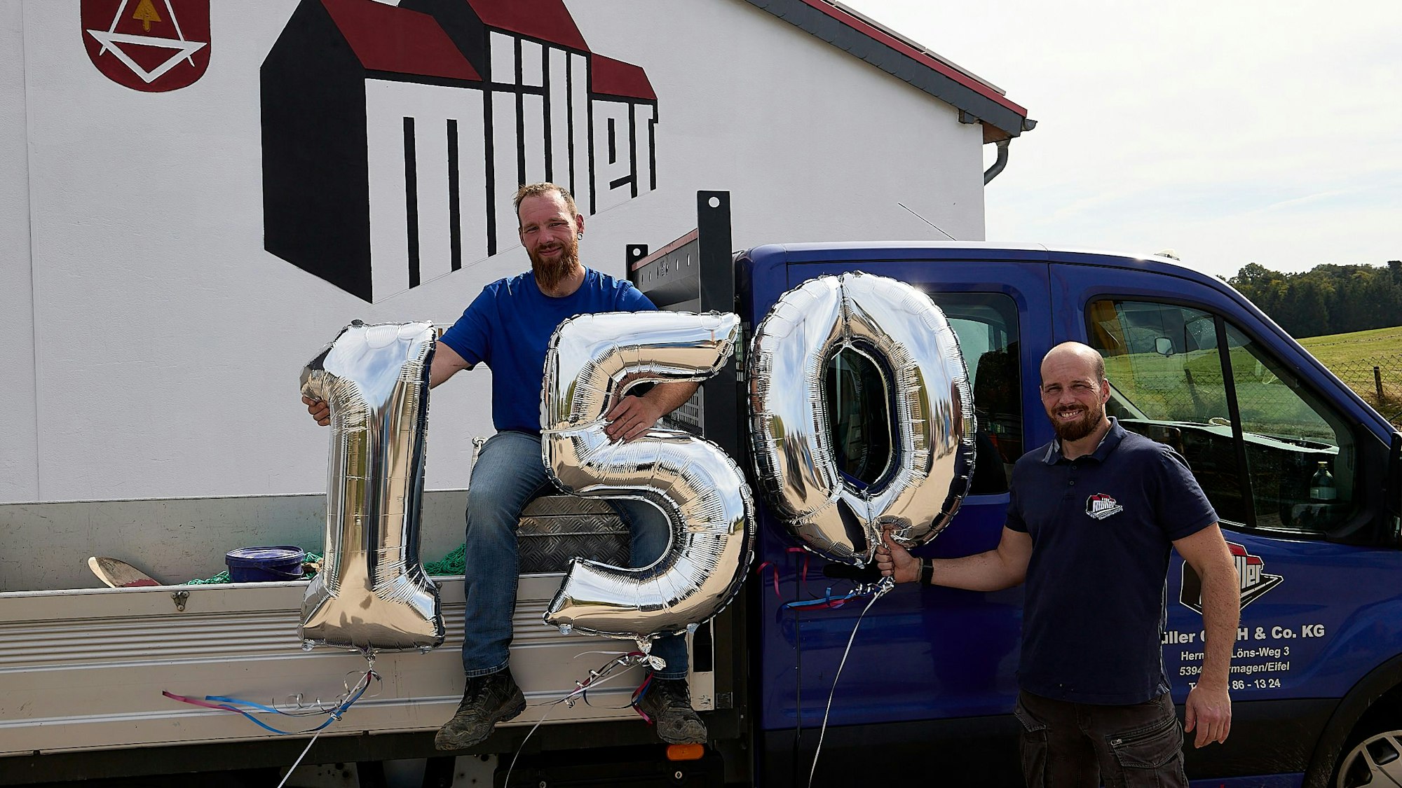 Ein Mann sitzt auf einem Lastwagen, ein anderer steht neben der Beifahrertür. Sie halten drei Luftballons, die die Zahlen 1, 5 und 0 bilden.
