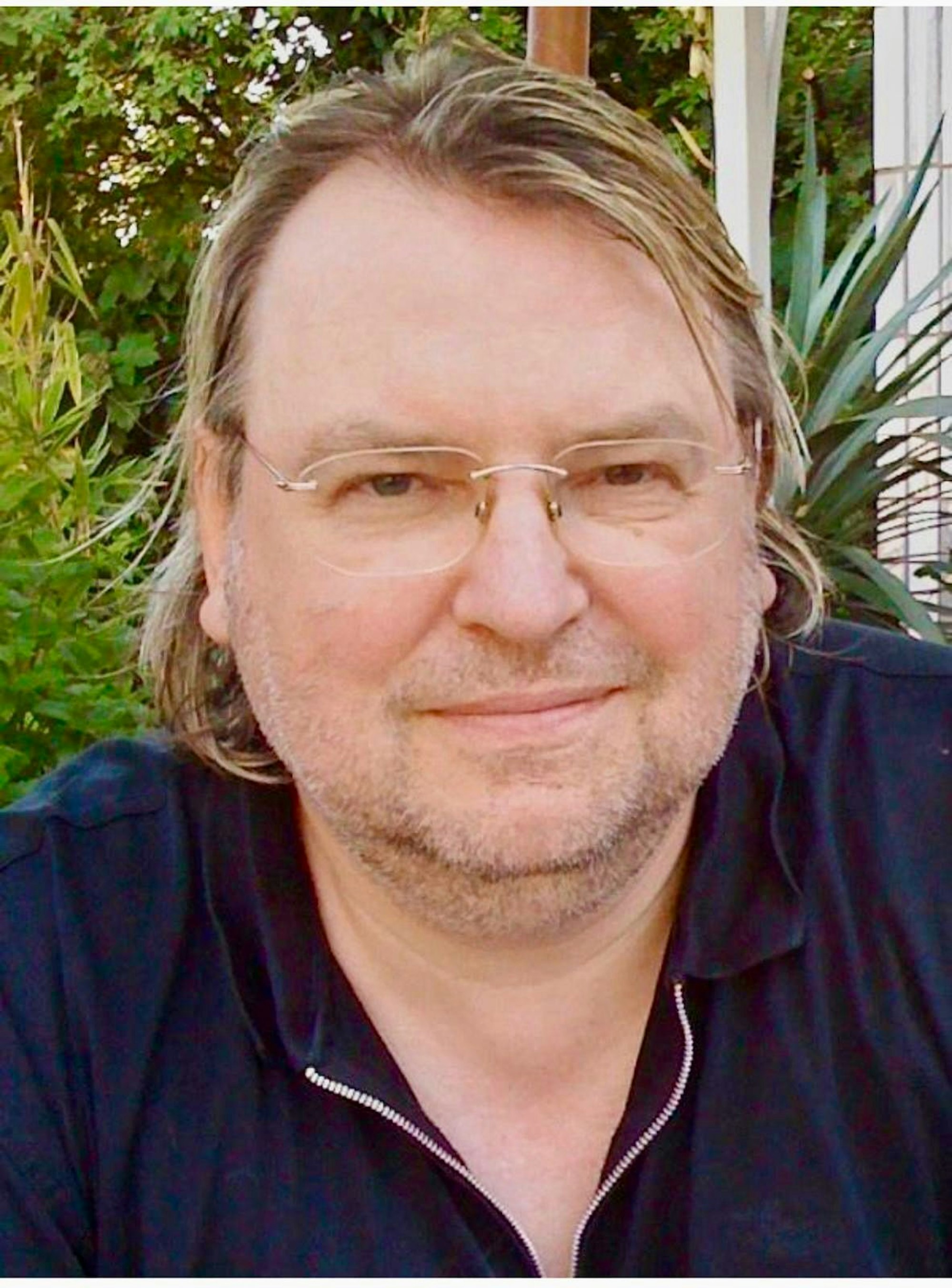 Der Komponist trägt eine randlose Brille und hat schulterlange blonde Haare und einen Dreitagebart.