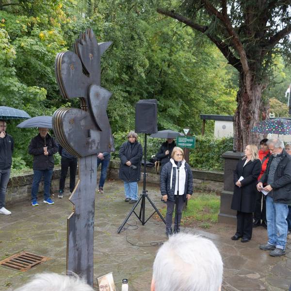 Eine Gruppe von Menschen steht im Halbkreis um eine aus Cortenstahl gefertigte Skulptur herum. Einige haben Regenschirme aufgespannt.