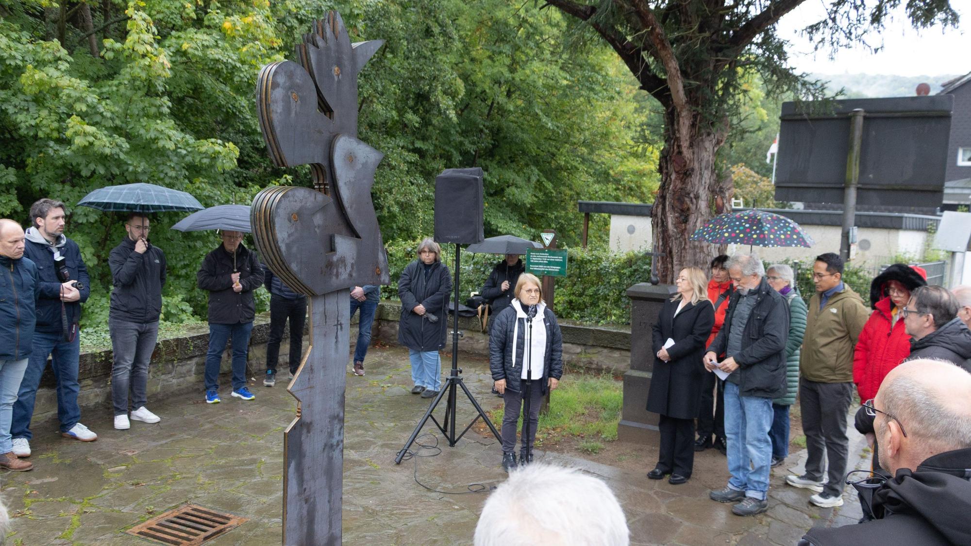 Eine Gruppe von Menschen steht im Halbkreis um eine aus Cortenstahl gefertigte Skulptur herum. Einige haben Regenschirme aufgespannt.
