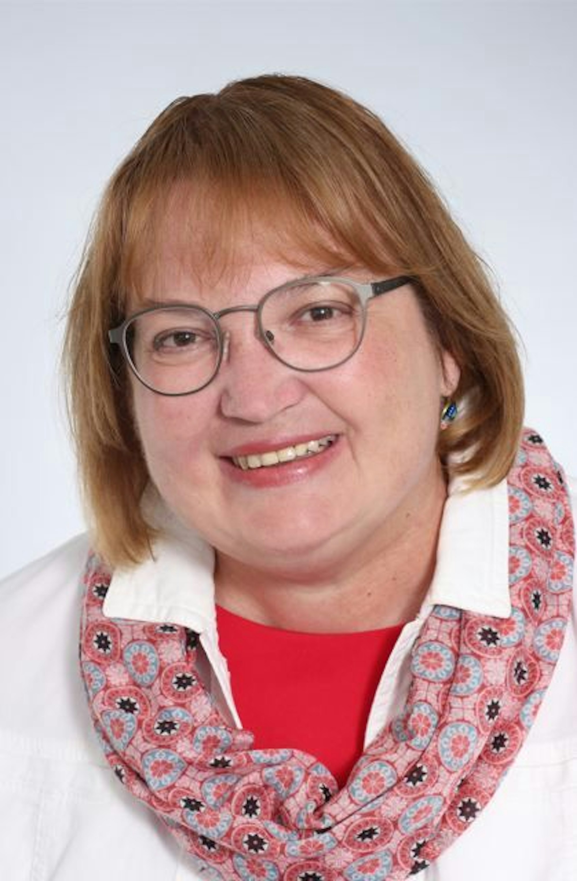 Das Bild zeigt Astrid Machuj. Sie hat schulterlanger glatte Haare und trägt eine Brille. Sie trägt ein weißes Hemd und einen gemusterten rötlichen Schal.