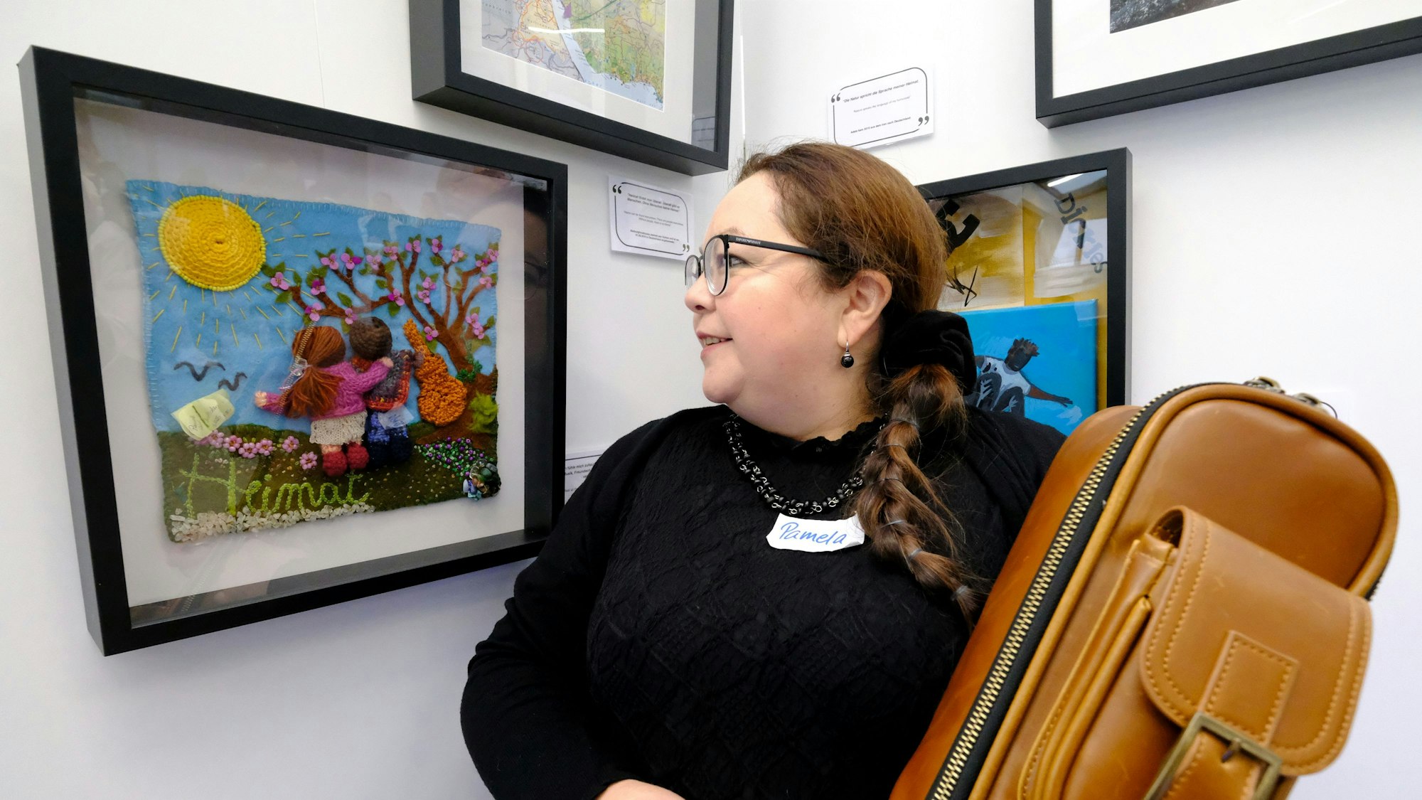 Eine Frau mit Brille und schwarzem Pullover blickt auf ein gerahmtes Kunstwerk, das mit anderen Arbeiten in einer Ausstellung zu sehen ist.