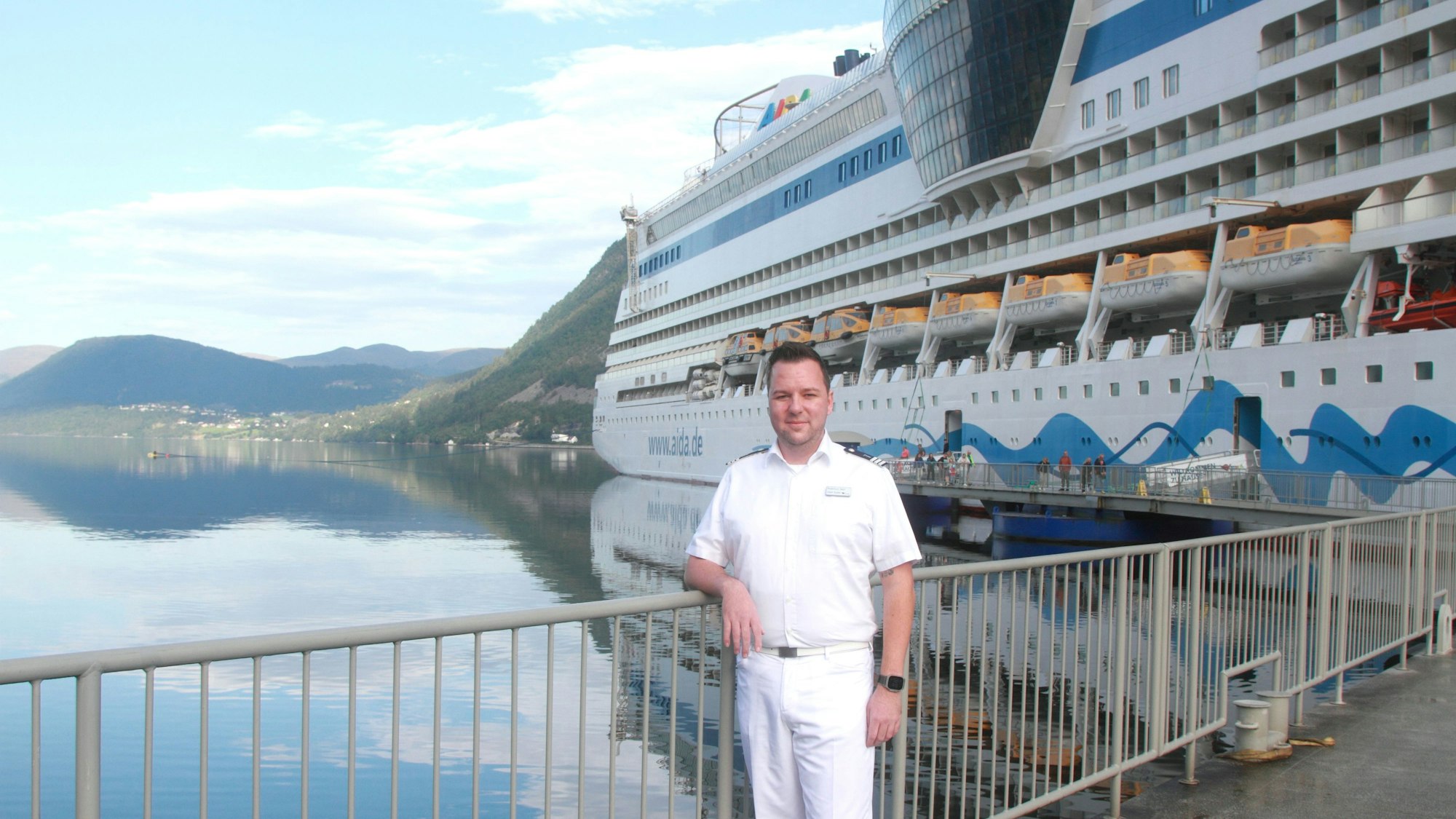 Federico Sass aus Euskirchen ist Chief Purser der Aida-Kreuzfahrtflotte. Hier steht er in seiner weißen Dienstkleidung vor einem Schiff.