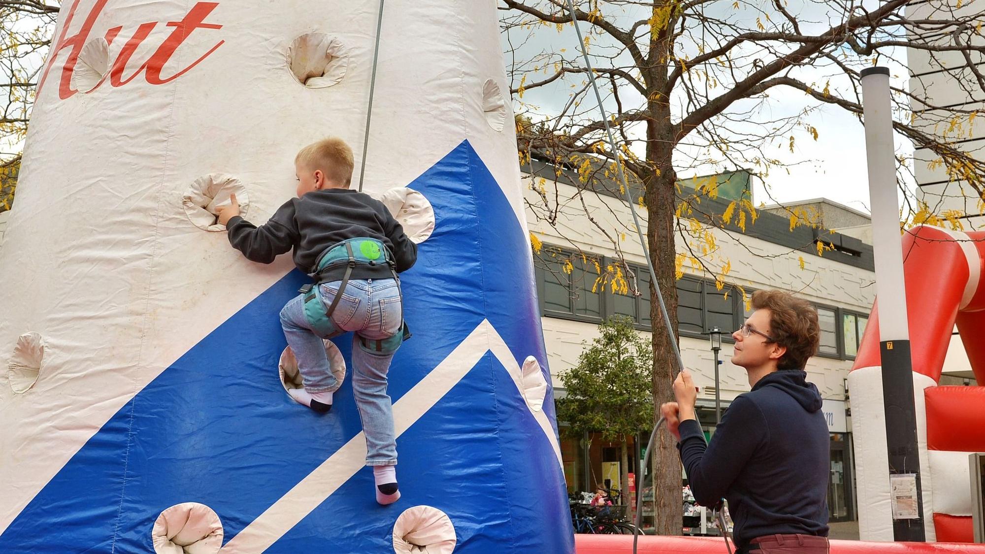 Ein junge klettert eine an einer Kletterwand. Daneben steht ein Mann, der ihn mit einem Kletterseil sichert.