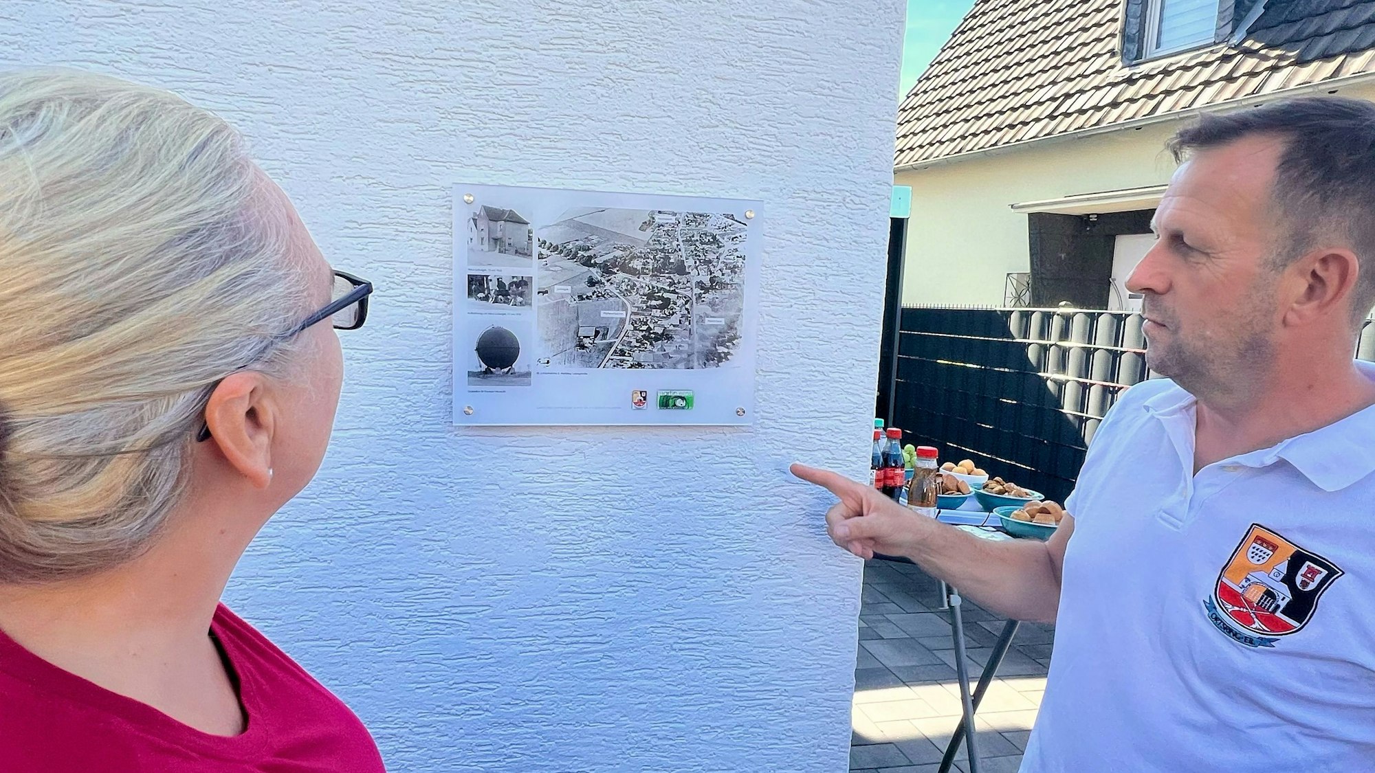 Ein Mann und eine Frau betrachten eine Fototafel, die an einer Hauswand angebracht ist.