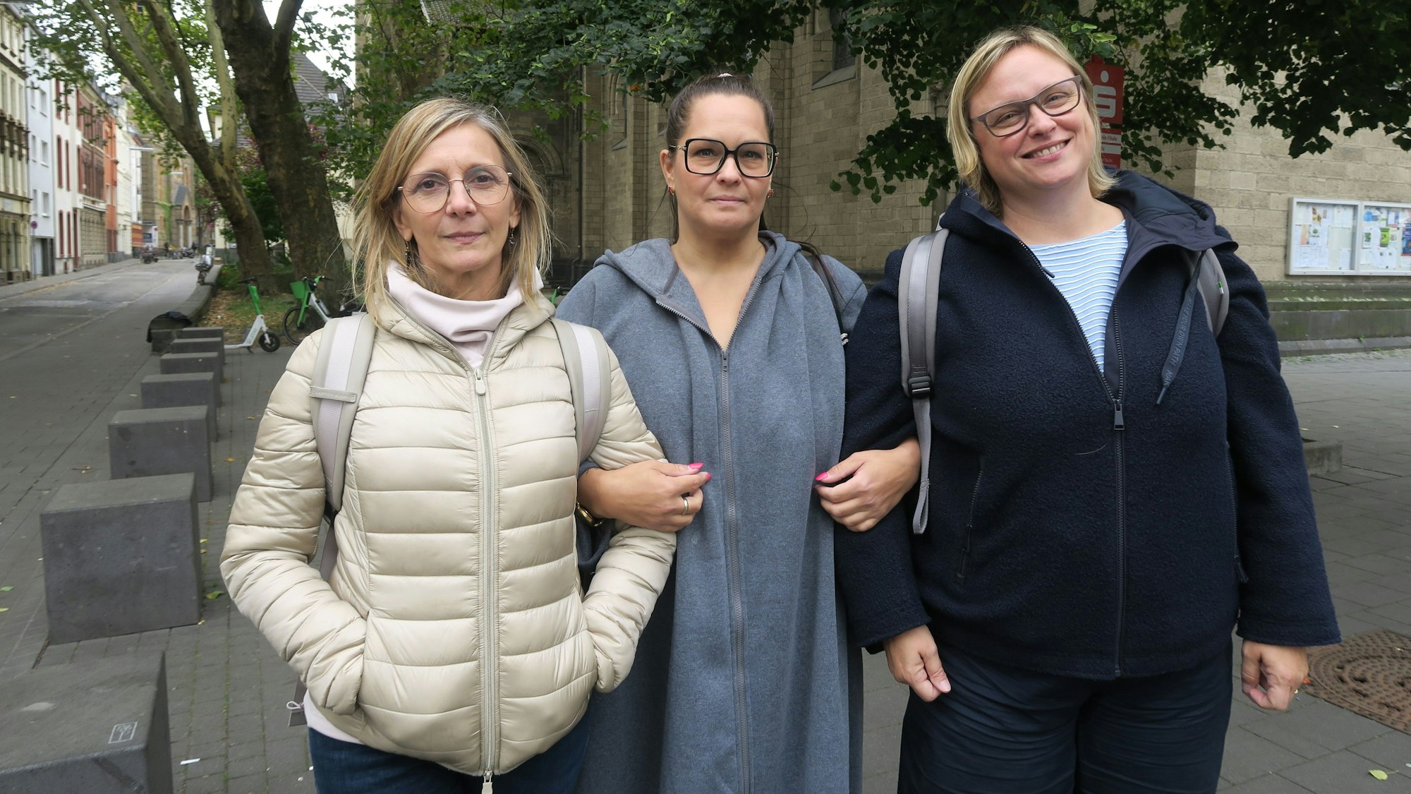 Drei Frauen stehen untergehakt auf einem Platz vor einer Kirche.